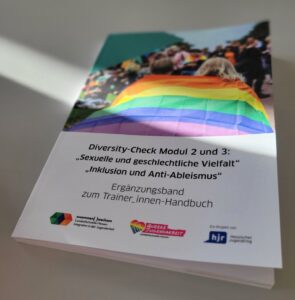 Auf dem Foto ist eine Ausgabe des Diversity-Check Handbuchs zu sehen. Das Buch liegt auf einem Tisch, die Sonne scheint darauf.