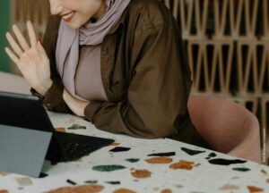 Auf dem Bild ist eine Person zu sehen, die vor einem Laptop sitzt, lächelt und winkt.